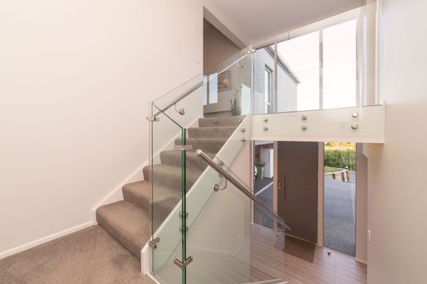 Bright contemporary residential stairway with carpeted stairs, clear glass balustrades, polished metal handrails and fittings, and a white structural landing beam. A tall glazed entry area and timber-toned flooring are visible beyond the stairs.