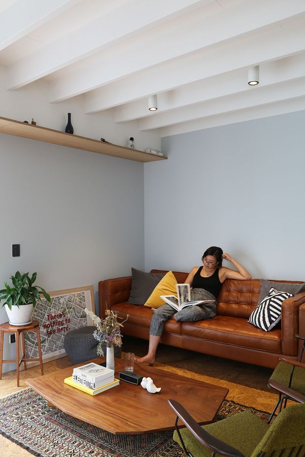 Bright, inviting living room with white exposed ceiling beams, pale blue-grey walls, a long timber display shelf, and a cognac leather sofa. The room includes mid-century-inspired seating, a large patterned rug, a wood coffee table, framed artwork, plants, books, and decorative objects. A seated person is reading on the sofa.