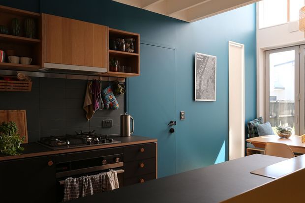 A compact, richly coloured kitchen with dark charcoal lower cabinetry and benchtops, warm timber overhead cupboards, open shelving, and a dark tiled splashback. Deep teal-blue walls create a strong feature colour, while a bright glazed dining area with timber furniture is visible to the right. The room has abundant natural light and a partially vaulted or raised ceiling with exposed beam-like detailing.