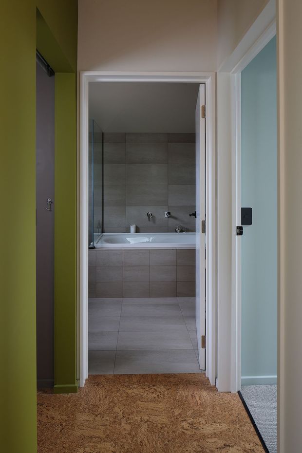 View from a hallway into a modern tiled bathroom. The bathroom contains a built-in bathtub with a tiled apron, grey large-format wall and floor tiles, and a clear glass shower screen. White door and trim frame the opening, with green-painted hallway walls and cork-like brown flooring in the foreground.