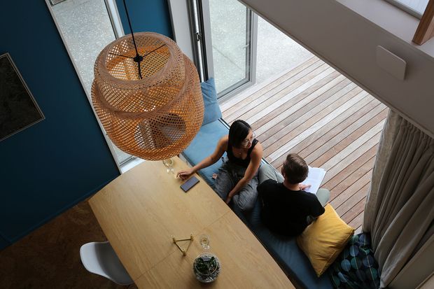 Overhead view of a contemporary dining and living area beside full-height glazing and a timber deck. A large light-wood dining table with white seating occupies the foreground, while a built-in window seat with cushions and curtains provides informal lounge seating. A woven pendant light hangs above the table, and a deep blue feature wall adds contrast.