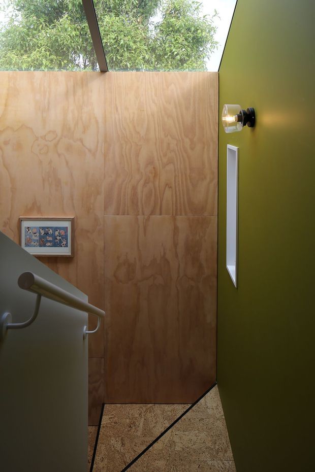 A compact, visually striking stairway with plywood wall panelling, a dark olive-green wall, a narrow vertical window, and a skylight overlooking leafy trees. The stair has a simple metal handrail, patterned carpeted treads, and a small framed artwork. Natural light and contrasting timber and green surfaces create a warm yet bold interior.