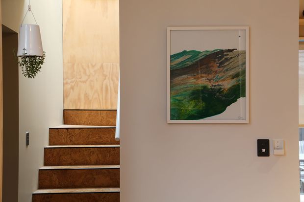 Interior view of a compact stairway with timber-toned stair treads and pale walls. A hanging pendant planter is positioned beside the stairs, while framed abstract artwork and wall switches decorate the adjacent wall. A glimpse of another room is visible through the opening at right.