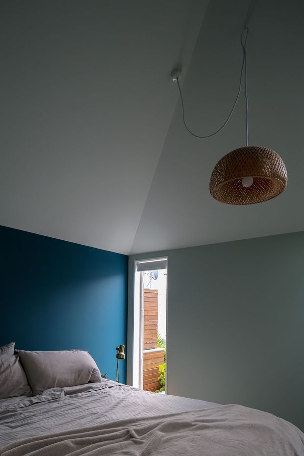 A moody bedroom interior with a low bed dressed in rumpled light-grey bedding, a deep blue accent wall behind the bed, and pale grey-green walls. A woven rattan pendant light hangs from the angled ceiling, while a narrow window provides a glimpse of timber screening and greenery outside. A small brass reading lamp stands beside the bed.