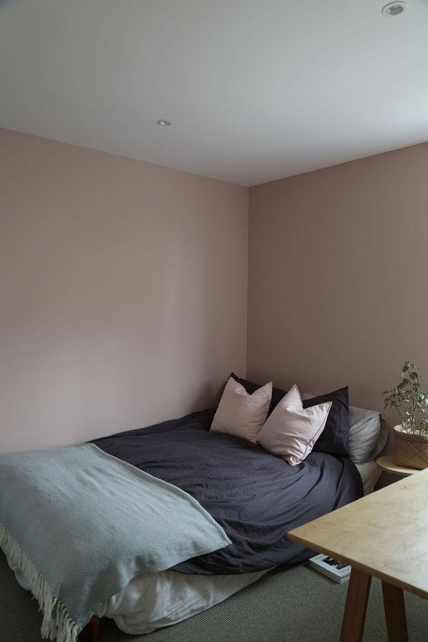 A compact bedroom with dusty rose walls, a low bed dressed in dark charcoal bedding and pale blush pillows, and a light blue-green fringed throw. A small potted plant sits beside the bed, while a simple light timber table occupies the foreground. Recessed ceiling lights and carpet flooring complete the understated, cozy interior.