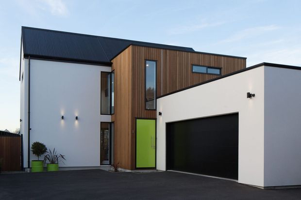 Contemporary multi-storey residential facade with white rendered walls, a dark pitched metal roof, vertical timber cladding, tall narrow windows, a vivid lime-green entry door, and an integrated black double garage. The building fronts a paved driveway and has a clean, geometric composition.