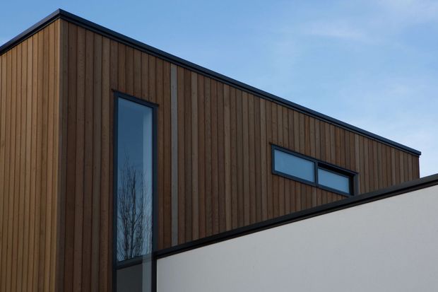 Exterior architectural view of a contemporary building with vertically clad timber walls, crisp black window and roof trims, and a contrasting smooth white wall in the foreground. Narrow vertical and horizontal windows create a clean, minimalist facade composition.