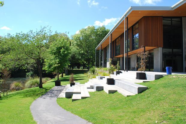 Contemporary single-storey residence set within a landscaped garden, viewed from an outdoor path. The building features extensive floor-to-ceiling glazing, warm vertical timber wall cladding and soffits, exposed concrete elements, a low-pitched roof with broad eaves, and stepped concrete terraces leading to the lawn. Mature trees and a small pond-like water feature surround the home.