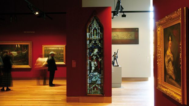 Interior view of a gallery or museum exhibition space with richly painted deep-red walls, polished timber flooring, framed artworks, a prominent Gothic-style stained-glass window panel, ornate gilded picture frames, track lighting, and visitors viewing the exhibits. A brighter adjoining gallery is visible through the opening.