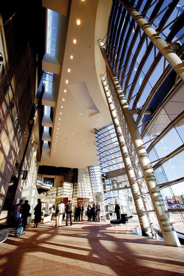 A tall, sunlit contemporary commercial atrium with a dramatic glazed curtain-wall roof and façade, exposed structural steel columns, and multiple suspended or cantilevered floor levels. A broad tiled concourse occupies the foreground, with people, seating, and retail or public-space activity below a large white soffit fitted with recessed downlights. Strong daylight creates long shadows across the floor, emphasizing the generous vertical scale and indoor-outdoor visual connection.