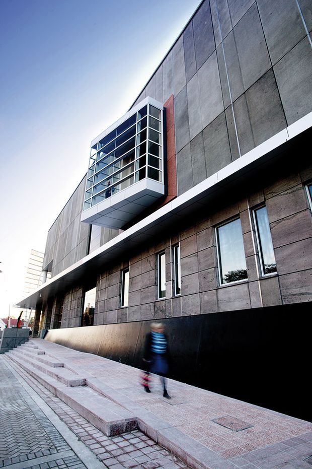 Exterior street-level view of a contemporary multi-storey building with a large exposed concrete panel facade, dark base, projecting glazed balcony or enclosed terrace with white metal framing, and narrow vertical windows. A stepped paved pedestrian walkway runs alongside the building.