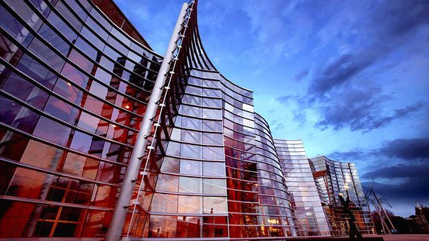 Low-angle exterior view of a large contemporary commercial building with sweeping curved glass curtain walls, reflective glazing, exposed structural elements, and red-toned interior or facade accents. The building forms a dramatic multi-storey urban architectural composition against a blue evening sky.