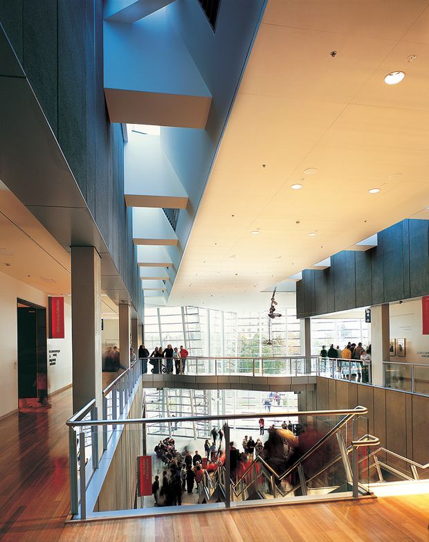 A large multi-level public interior atrium with broad timber stair landings, metal-and-glass balustrades, extensive floor-to-ceiling glazing, suspended sculptural elements, and crowds gathered across the lower levels. The space has a contemporary institutional or commercial character, with recessed ceiling lighting and dark textured wall panels.