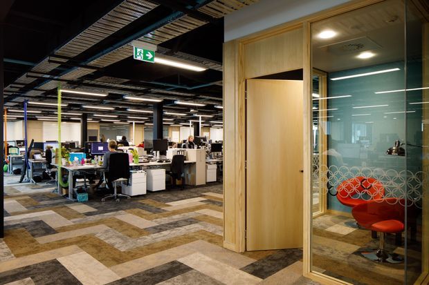 Open-plan contemporary office interior with exposed dark ceiling services, rows of workstations, patterned carpet tiles, and a timber-framed glazed meeting or breakout room. The glass room contains bright red lounge seating and decorative circular privacy film.