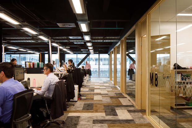 Open-plan commercial office interior with rows of workstations on the left, glazed meeting rooms and timber-framed partitions on the right, and a broad circulation corridor through the centre. The space has an exposed dark ceiling with visible services, linear fluorescent lighting, patterned carpet tiles, and a contemporary workplace fit-out.