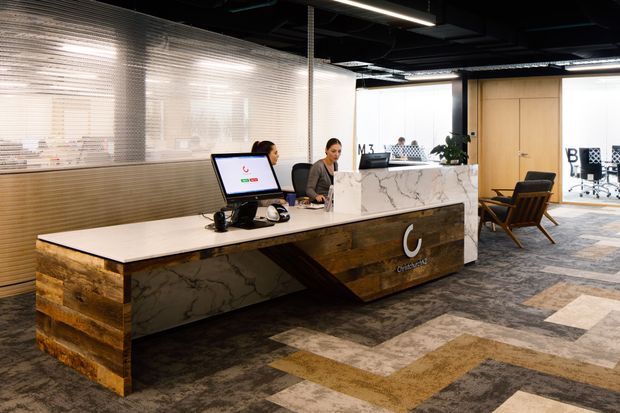 A contemporary commercial reception area with a large sculptural reception desk featuring a white marble-look surface and warm reclaimed timber cladding. The desk includes a prominent illuminated logo, computer workstation, and reception staff. Behind it are translucent textured glass partitions, timber doors, and glazed meeting or office spaces. The interior has an exposed black ceiling with linear lighting and a patterned grey, beige, and brown carpeted floor. Lounge chairs and indoor planting add seating and softness.