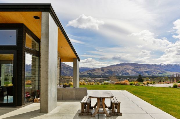 A contemporary single-storey home opens onto a broad paved terrace with a timber picnic table and built-in outdoor seating. The facade combines dark-framed floor-to-ceiling glazing, textured dark cladding, pale rendered structural columns, and a warm timber-lined soffit. Beyond the manicured lawn is an expansive mountain landscape under a partly cloudy sky.