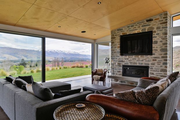 A spacious contemporary living room with floor-to-ceiling corner glazing overlooking a landscaped lawn, vineyard-like planting and mountain scenery. The room features a large grey sectional sofa, leather seating, a stone-clad fireplace wall with built-in fireboxes, and a warm timber-lined ceiling with recessed lighting.