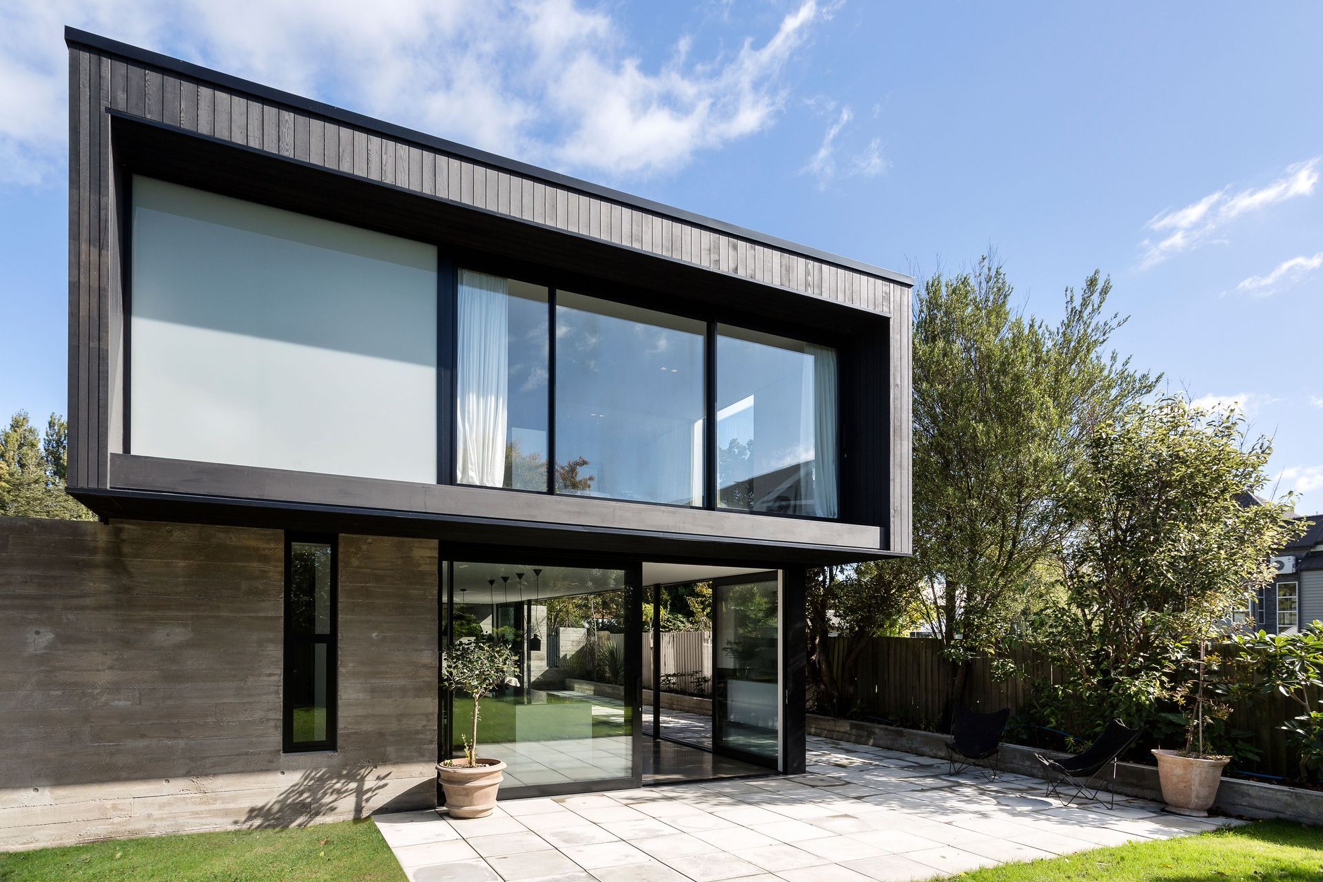 The view from the back garden shows the upper box perched over the concrete wall and the glazed living area below. Upstairs, the master bedroom overlooks mature trees and the garden.