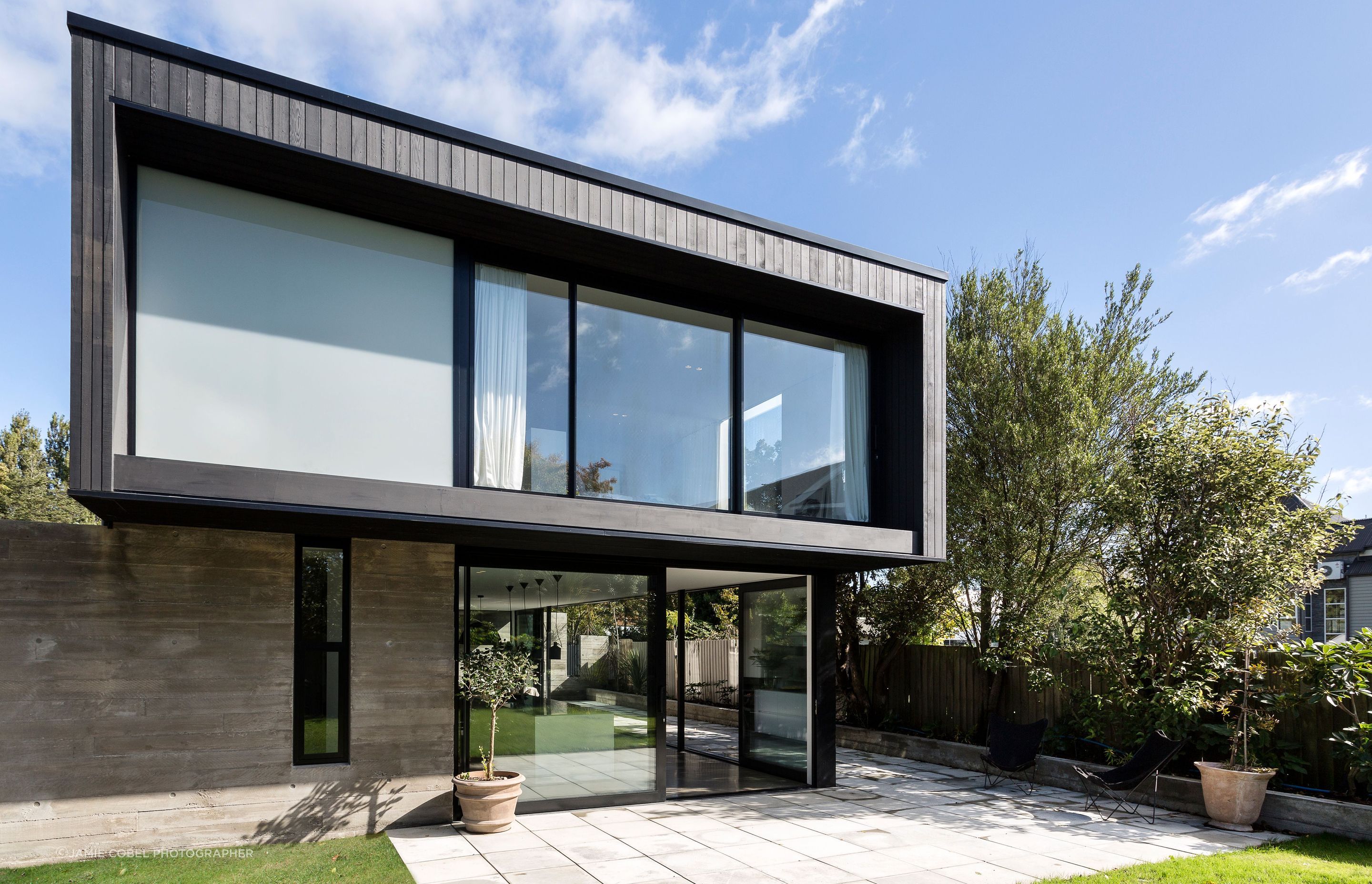 The view from the back garden shows the upper box perched over the concrete wall and the glazed living area below. Upstairs, the master bedroom overlooks mature trees and the garden.