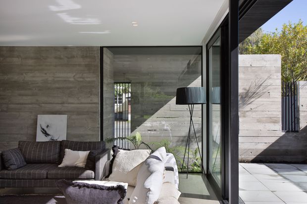 A contemporary living room opening onto a paved courtyard through expansive black-framed glazing. The room features a dark upholstered sofa, light lounge seating, a floor lamp, concrete-look wall finishes, and a smooth white ceiling. Strong daylight and the indoor-outdoor connection create a refined, modern atmosphere.
