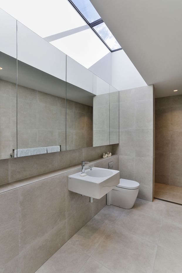 A bright, contemporary bathroom with large-format beige-grey porcelain tiles covering the floor and walls. A wall-mounted white basin and toilet sit beneath a large frameless mirrored wall, while a glazed skylight brings in abundant natural light. A walk-in shower area is visible at the right.