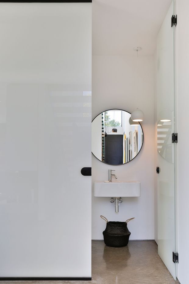 A compact minimalist bathroom viewed through a partially open white door. The space features a wall-mounted white basin with exposed chrome plumbing, a round black-framed mirror, a clear glass shower enclosure, white walls, and a polished grey concrete-like floor. A black woven basket sits beneath the basin, while pendant-style wall lighting and reflected linear details add visual interest.