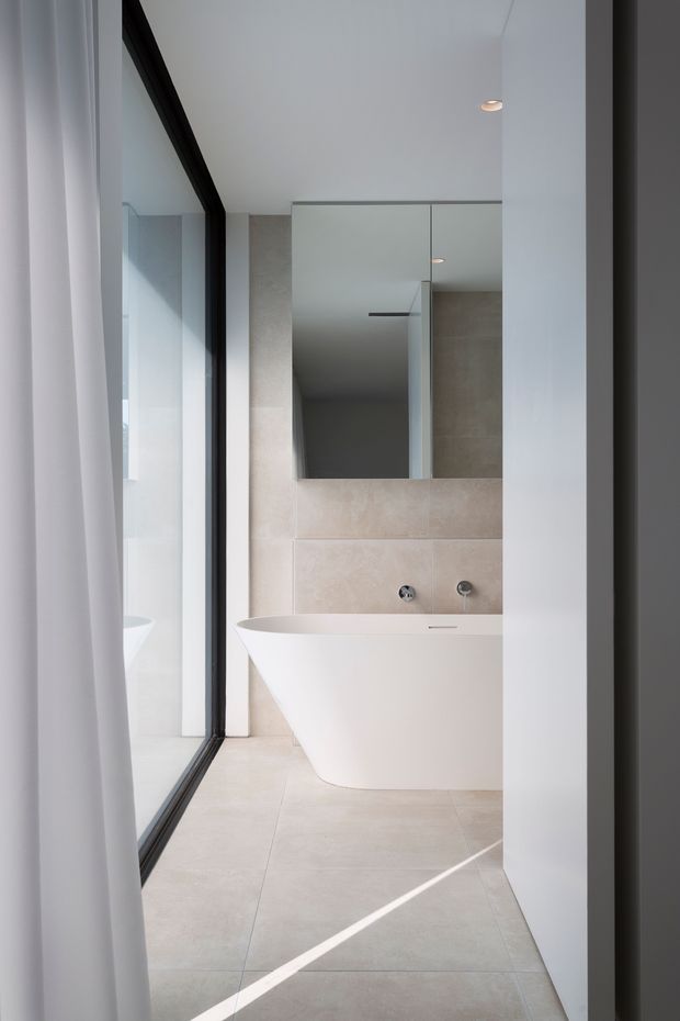 A bright, minimalist bathroom viewed through a doorway, featuring a freestanding white bathtub, large mirrored wall cabinetry, pale beige stone-look wall and floor tiles, and a full-height black-framed window or glazed opening. Clean recessed lighting and restrained detailing create a refined contemporary interior.