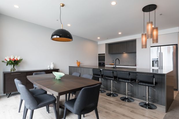 Contemporary open-plan kitchen and dining area with a dark wood dining table, upholstered dining chairs, a long island with black bar stools, dark wood cabinetry, grey tiled backsplash, stainless-steel appliances, and layered pendant lighting. The room has light wood-look flooring, pale grey walls, and a neutral, refined palette.