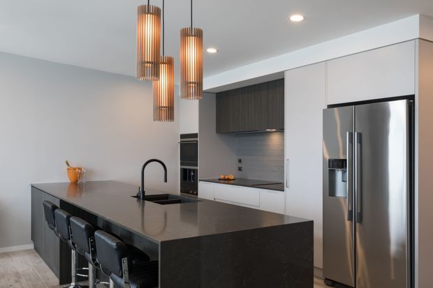 Contemporary kitchen with a large dark stone island featuring an integrated sink and curved black tap, seating for four at bar stools, white full-height cabinetry, dark wood overhead joinery, a textured light-grey splashback, and stainless-steel appliances. Three linear pendant lights add a warm accent above the island.