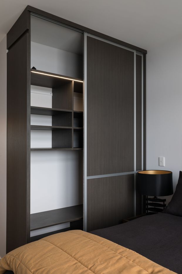 A contemporary bedroom featuring a full-height dark wood built-in wardrobe with sliding doors, open shelving, integrated LED lighting, and hanging storage. A dark bed with a mustard-brown quilt and a black bedside lamp occupies the foreground. The room has plain light walls and a restrained, dark-toned palette.