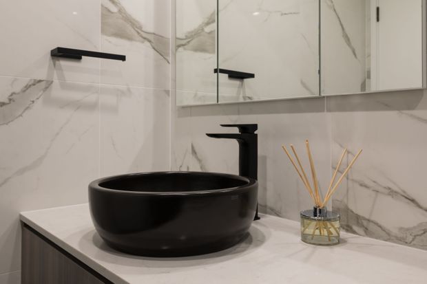 A contemporary bathroom vanity featuring a glossy black vessel basin, matte black wall-mounted tapware, white marble-look wall tiles with grey veining, and a mirrored wall cabinet. A reed diffuser sits on the pale stone-look benchtop.