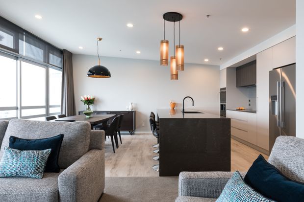Open-plan contemporary apartment interior combining a kitchen, dining area and living room. A dark stone island with waterfall edge and integrated sink anchors the kitchen, with white and dark cabinetry, stainless-steel appliances and pendant lighting. The dining area sits beside floor-to-ceiling glazing, while grey upholstered sofas and patterned cushions furnish the living zone. Light timber-look flooring and neutral walls create a bright, refined space.