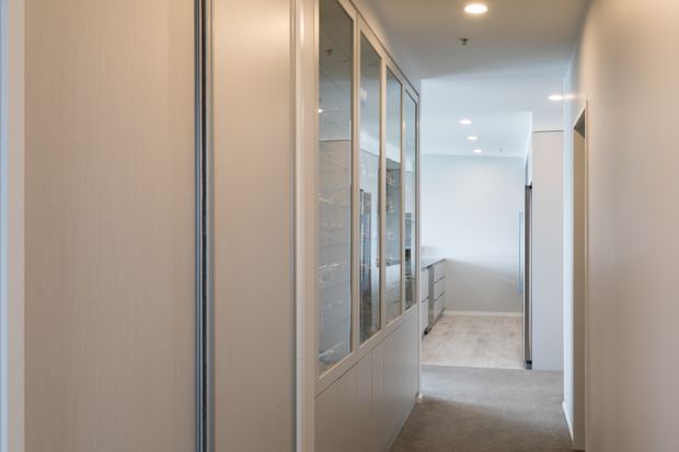 A bright, narrow interior hallway with pale carpet, white walls, recessed ceiling lights, and light-toned built-in cabinetry. A long glazed display or storage partition with slim metallic frames lines the left side, while the corridor opens toward a stair or landing area at the far end.