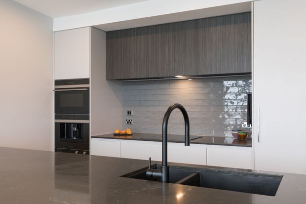 Modern kitchen featuring a large dark stone island with an integrated double-bowl sink and tall black gooseneck tap. White lower cabinetry and tall appliance housing contrast with dark vertically grained upper cabinetry. A glossy white subway-tile splashback is illuminated beneath the wall cabinets, creating a clean, refined contemporary fit-out.
