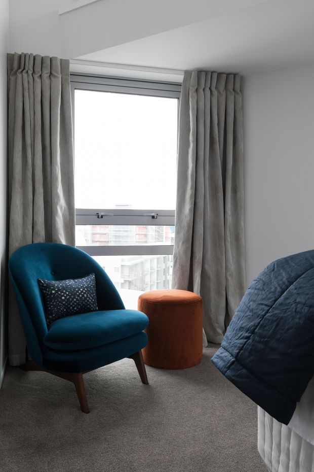 A softly lit bedroom corner with a large window framed by full-length pleated grey curtains. The room features a deep teal upholstered lounge chair with a patterned cushion, a rust-orange cylindrical ottoman, grey carpet, and a partially visible bed dressed in a dark blue quilt.