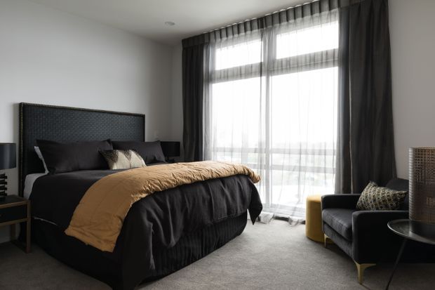 A sophisticated contemporary bedroom with a large upholstered bed dressed in black and charcoal bedding, accented by a warm mustard throw. Floor-to-ceiling sheer and dark curtains frame expansive glazing, while a dark armchair, side table, bedside tables, and layered lighting complete the room. The palette is moody and refined with soft natural light and plush textures.