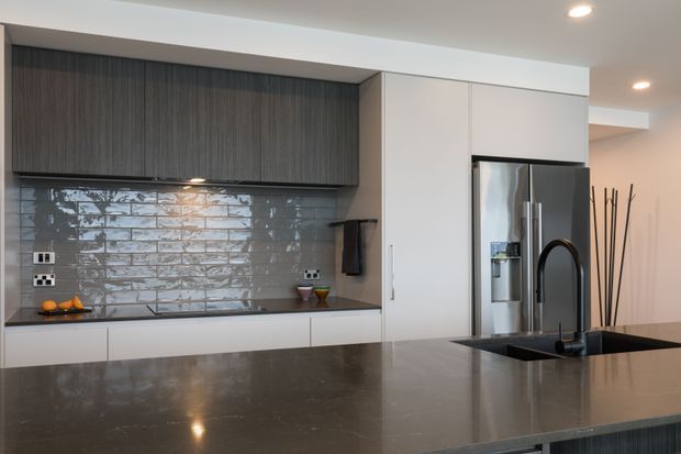 Contemporary kitchen with a large dark stone island and integrated black sink and gooseneck tap in the foreground. White full-height cabinetry surrounds a stainless-steel French-door refrigerator, while dark vertically grained overhead cabinetry contrasts with a glossy grey subway-tile splashback. Recessed ceiling lighting and a neutral palette create a refined, modern interior.