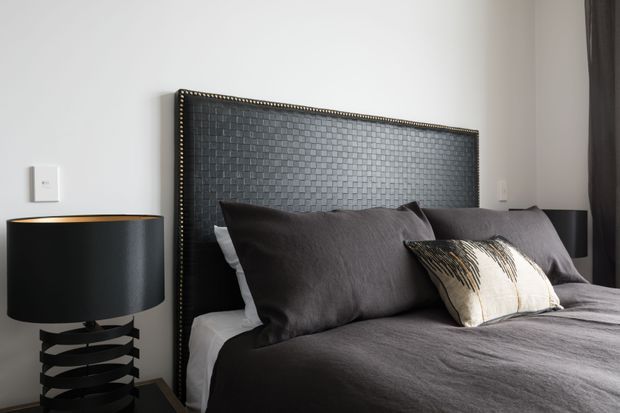 A close-up contemporary bedroom with a large upholstered charcoal-grey headboard featuring a woven, textured surface and nailhead trim. The bed is dressed in dark grey bedding with layered pillows, including a patterned accent cushion. Matching black bedside lamps sit on either side, with a white wall and dark curtain completing the restrained, monochromatic scheme.