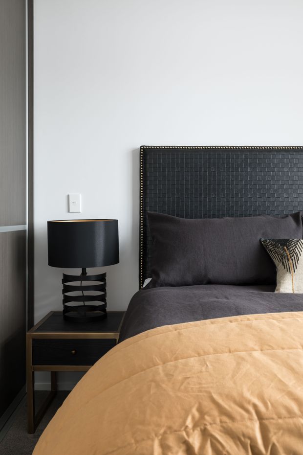 A close-up of a sophisticated bedroom featuring a dark upholstered bed with a textured, nailhead-trimmed headboard, charcoal bedding, and a warm tan duvet. A black bedside table with a sculptural black lamp sits beside the bed. The room has light grey walls, dark vertical wall panelling or cabinetry, and a restrained monochromatic palette with warm accents.