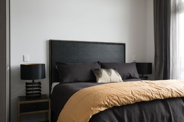 A contemporary bedroom featuring a dark upholstered bed with a tall textured headboard, charcoal bedding, and a warm tan throw. Matching black bedside tables and sculptural lamps flank the bed, with grey curtains and a large window on the right. The palette is dark, warm, and refined.