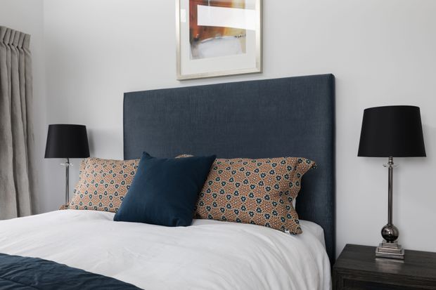 A styled bedroom featuring a large dark navy upholstered headboard, white bedding, patterned rust and blue pillows, matching black table lamps, dark bedside furniture, grey curtains, and framed abstract artwork.