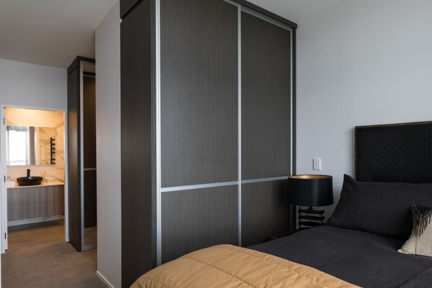 A contemporary bedroom with dark upholstered bedding, a tall black padded headboard, a black bedside table and lamp, and a large built-in dark wood wardrobe with sliding doors. An ensuite bathroom is visible through the open doorway, with a vanity and basin.
