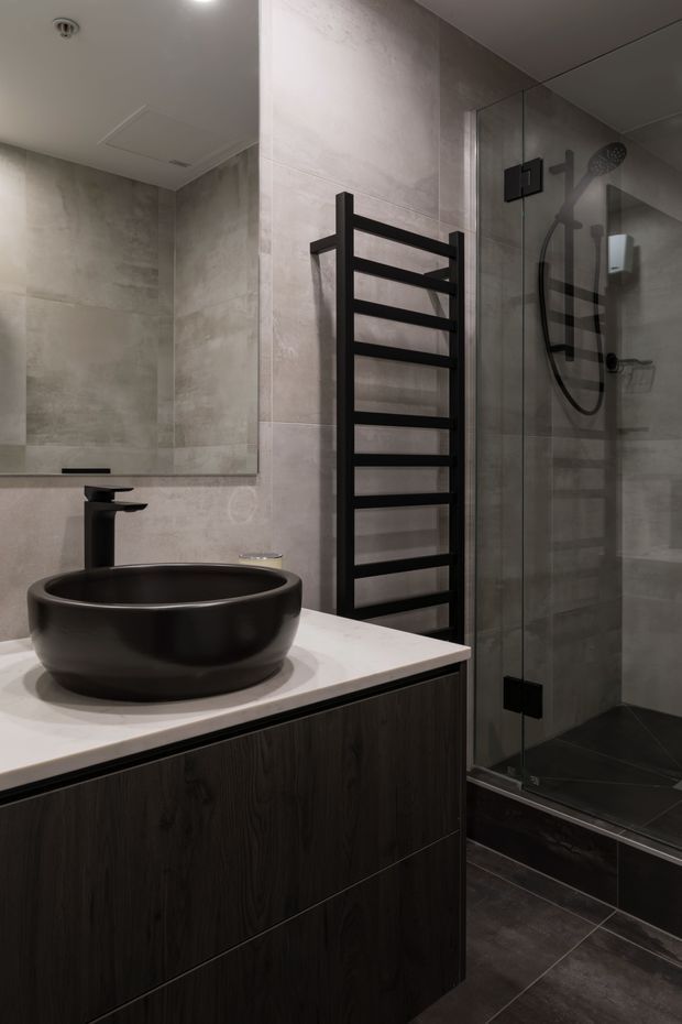 A contemporary bathroom featuring a black vessel basin on a white stone vanity top, dark timber-look drawer cabinetry, grey stone-look wall tiles, dark floor tiles, a black heated towel rail, and a frameless glass shower enclosure with matte black fixtures. The room has a refined, moody and high-contrast material palette.