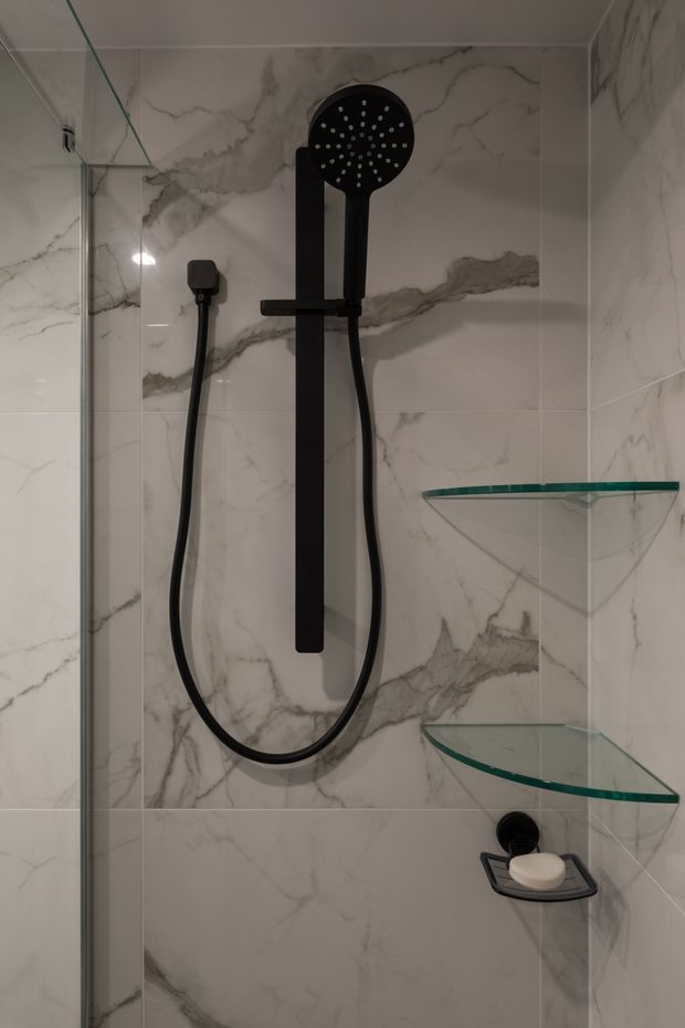 Close-up view of a contemporary shower enclosure with large white marble-look wall tiles featuring grey veining, matte black rain shower and handheld fixtures, frameless glass panels, and curved glass corner shelves with a black soap dish.