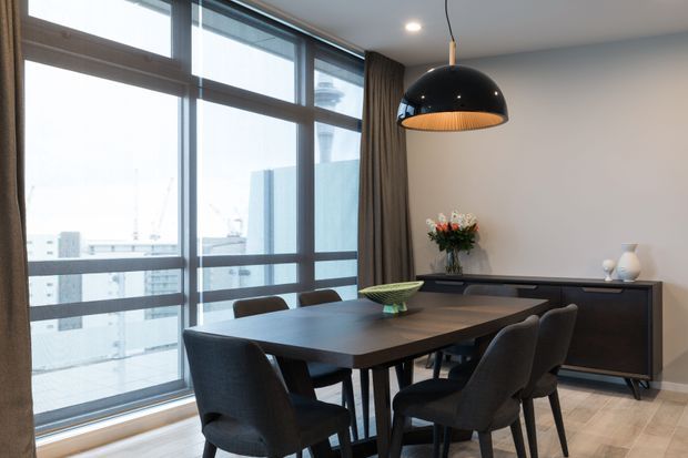 A contemporary dining area with a dark rectangular dining table surrounded by upholstered charcoal chairs. Floor-to-ceiling glazed windows and sliding doors line one side, with sheer and dark curtains, while a black dome pendant light hangs above the table. A dark timber sideboard with decorative vases and flowers sits against a neutral wall. Light timber flooring and restrained furnishings create a refined urban interior.