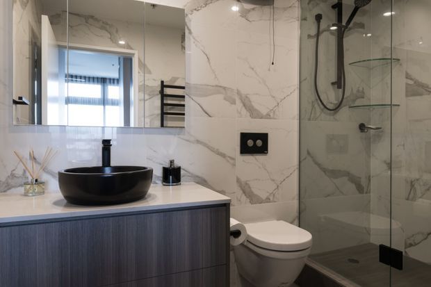 Contemporary bathroom with a floating dark wood-grain vanity, white benchtop, black vessel basin and tapware, wall-mounted toilet, and frameless glass shower enclosure. Large white marble-look wall tiles with grey veining create a polished, refined finish.