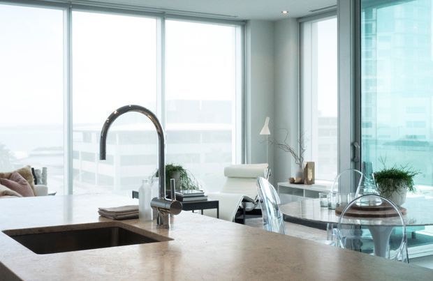 Bright contemporary open-plan kitchen overlooking a cityscape, with a large stone island and integrated sink, sculptural gooseneck tap, extensive floor-to-ceiling glazing, and a dining area with transparent acrylic chairs. Neutral finishes and abundant daylight create an airy, refined atmosphere.