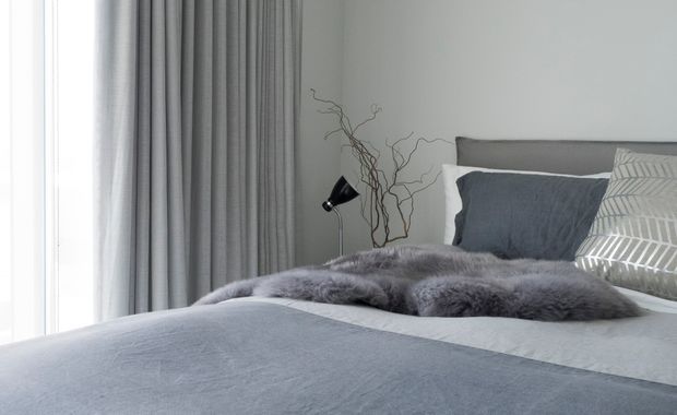 A softly styled contemporary bedroom featuring a low-profile upholstered bed, layered grey and white bedding, textured throw, geometric accent cushions, full-height grey curtains, and a black bedside lamp with decorative branches. The palette is calm and monochromatic with natural daylight entering from the left.