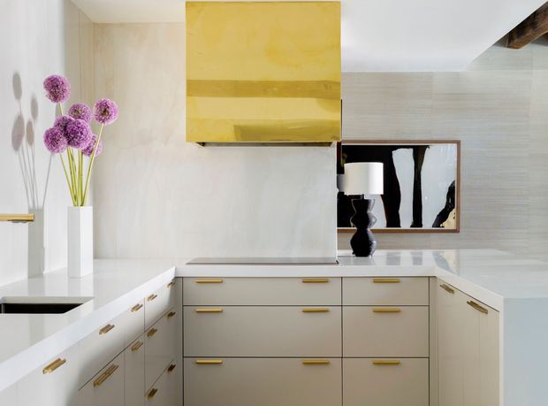 Bright, refined contemporary kitchen with a large U-shaped run of pale cabinetry and brass hardware, a light stone benchtop, and a concealed or integrated cooktop beneath a prominent yellow-gold rangehood. A white sculptural vase with purple flowers, a black decorative lamp, and framed artwork add accents. The room features pale textured wall finishes and a clean, tailored built-in design.